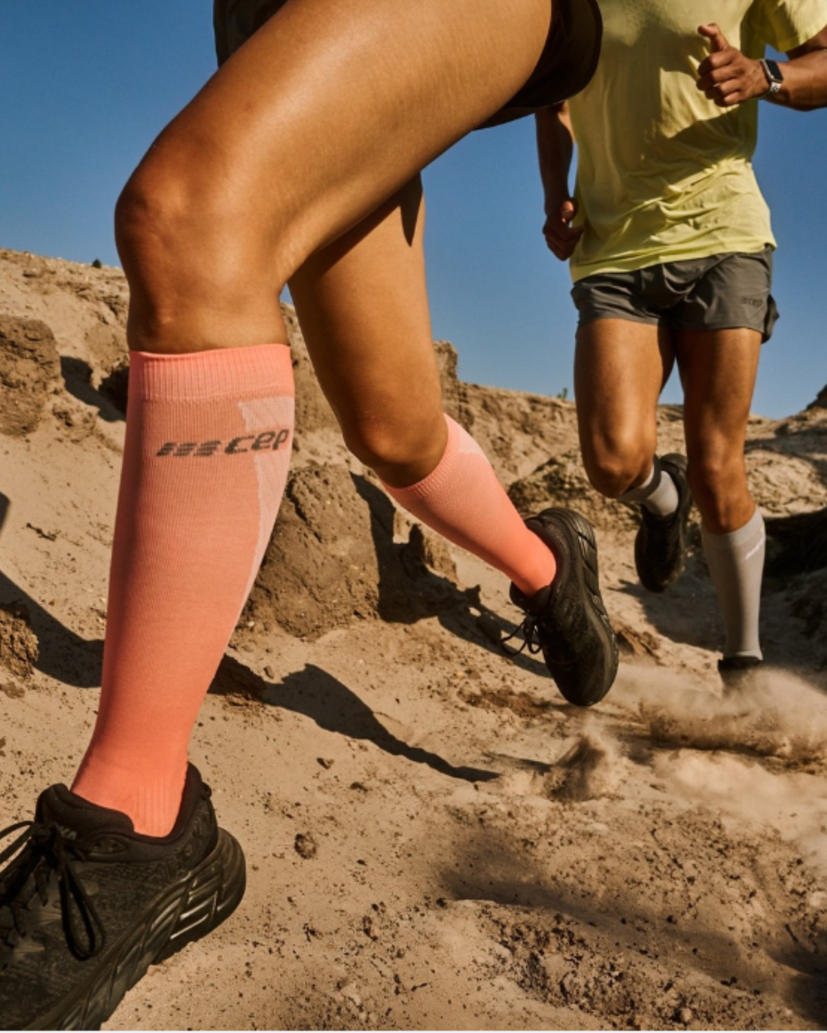 Compression socks in tall pink style worn by a woman trail running on dusty terrain alongside another runner