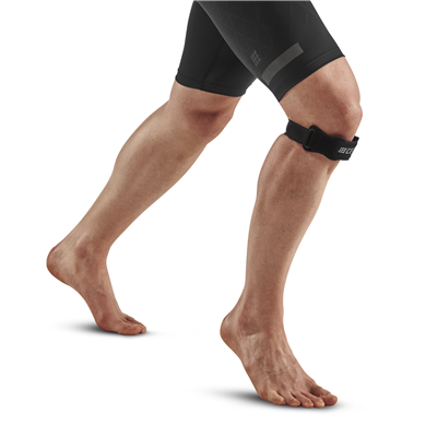 Patella strap in black worn just below the knee on a standing person's left leg, shown against a white background