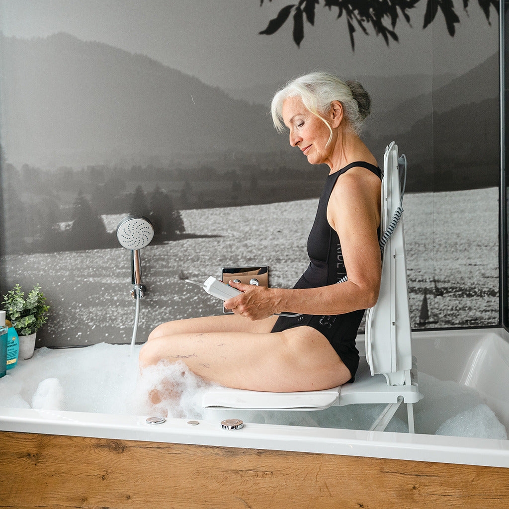 Bath lift with backrest in use inside a bathtub, shown with an older woman seated holding the handheld remote control