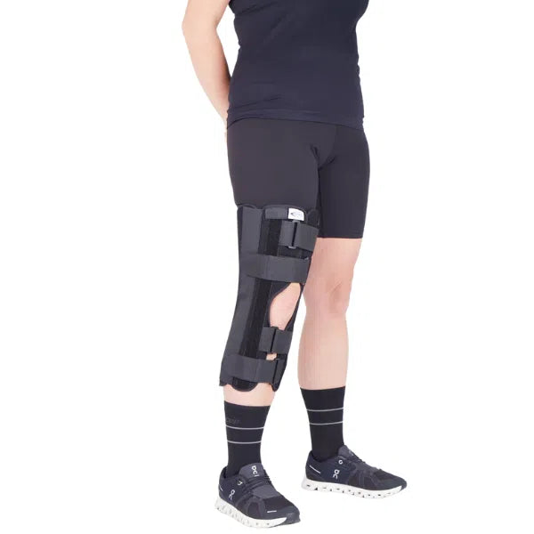 Knee immobilizer brace worn on left leg of person standing, black rigid panel design with hook-and-loop straps extending