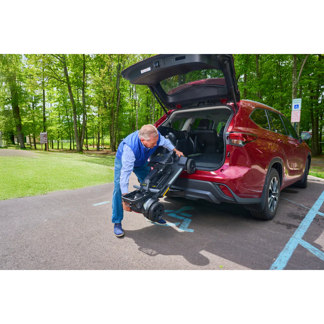 Folding travel scooter being loaded into the trunk of a red SUV by a man in a blue jacket in an accessible parking space