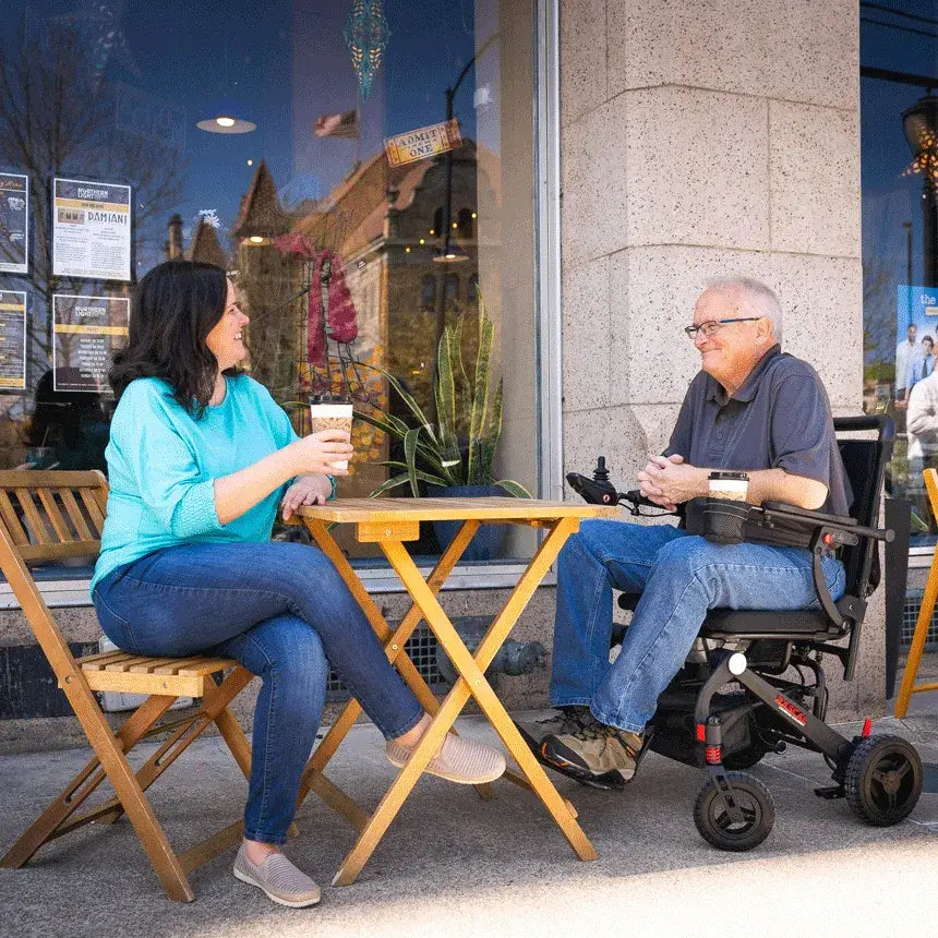 Power wheelchair user and companion enjoying coffee at an outdoor café table, with the Jazzy Rascal Carbon Cruiser parked