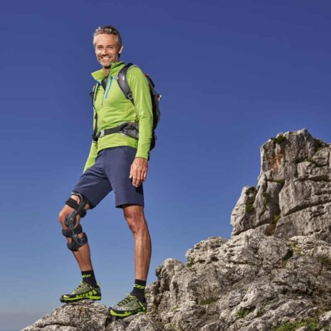 Knee brace worn by a smiling man standing on rocky mountain terrain with a backpack under a clear blue sky