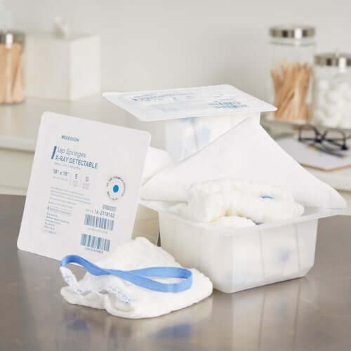 Surgical laparotomy sponges in open packaging with x-ray detectable label, blue loop ties, and bulk container on countertop