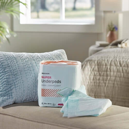 McKesson Super Underpads package with a single pad unfolded beside it, displayed on a beige sofa in a bedroom setting