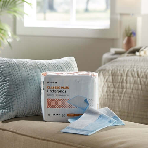 Disposable underpads package and two loose pads displayed on a beige sofa with a blue pillow and bedroom background