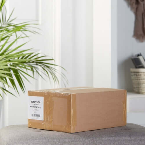 Disposable knit pants in sealed cardboard shipping box with McKesson label, resting on a gray upholstered surface