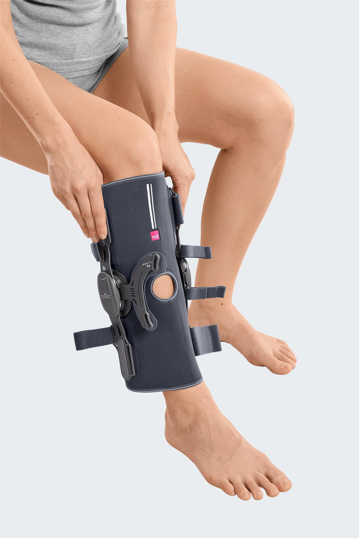 Knee brace with hinged side uprights, kneecap opening, and adjustable straps being fitted on a seated person's left leg