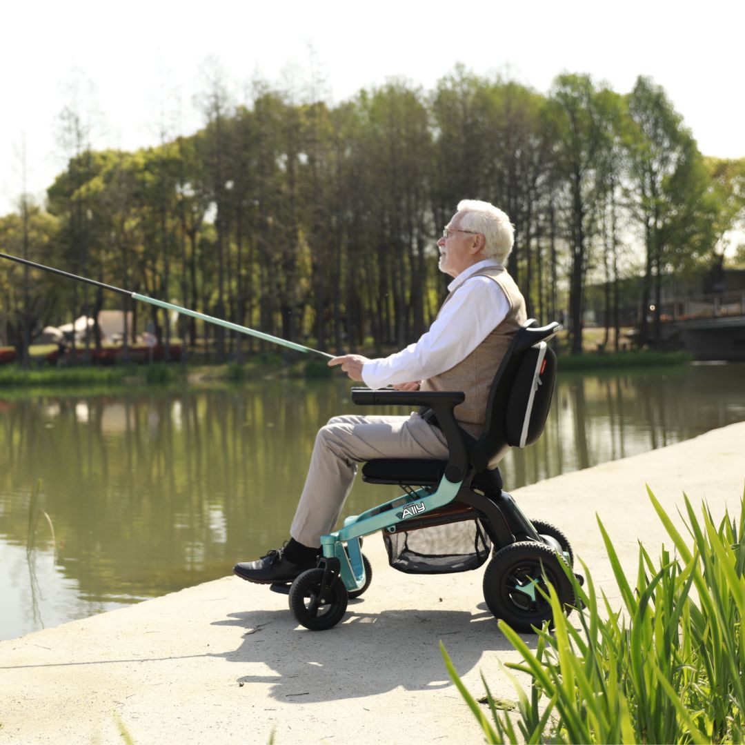 Power wheelchair with teal frame carrying an elderly man fishing beside a calm pond surrounded by trees
