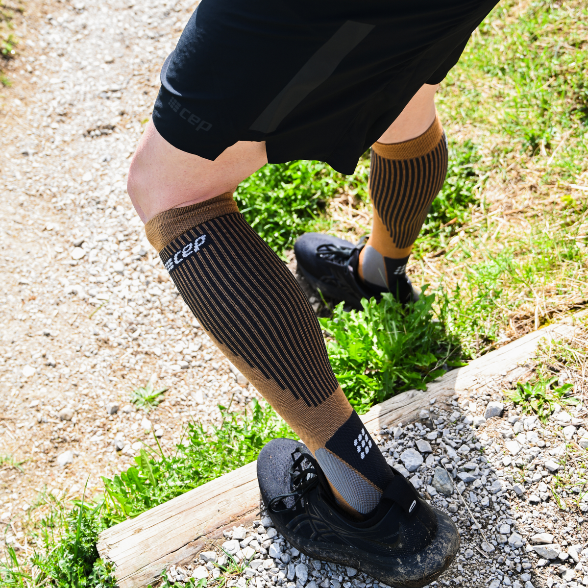 Compression calf sleeves in tan and black stripes worn over athletic socks on a woman's legs outdoors on a gravel trail