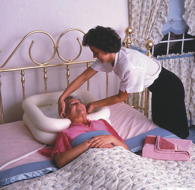 Inflatable shampoo basin cradling an elderly woman's head in bed while a caregiver washes her hair