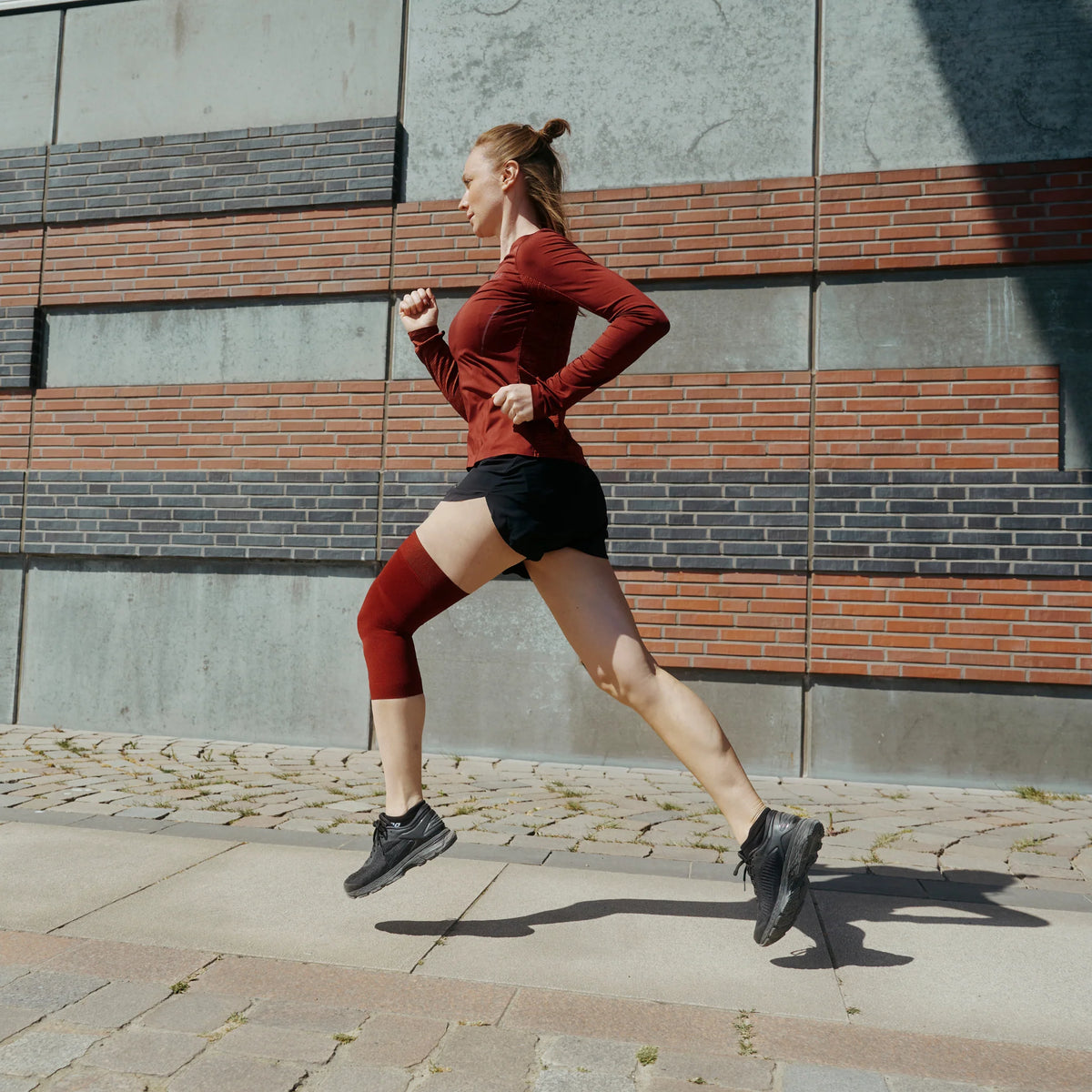 Knee sleeve in dark red worn on left knee of a woman running outdoors in athletic wear along a brick building