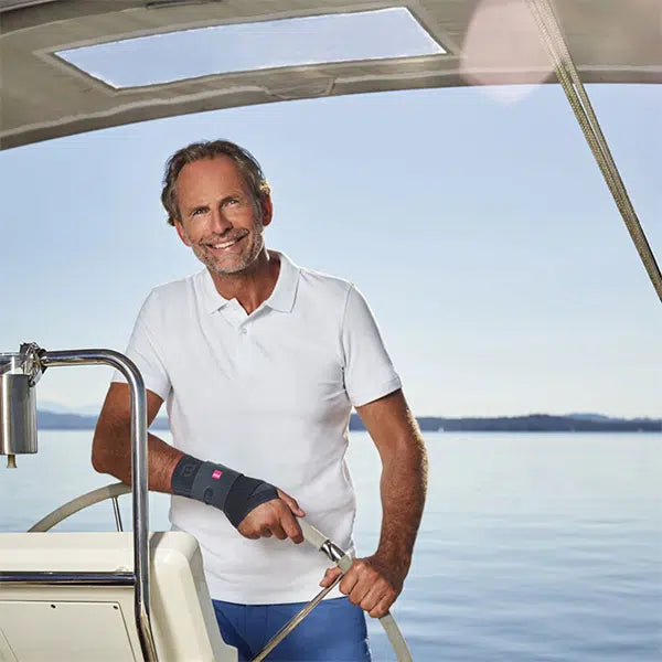 Wrist support worn by a smiling middle-aged man steering a boat on calm open water, dressed in a white polo shirt