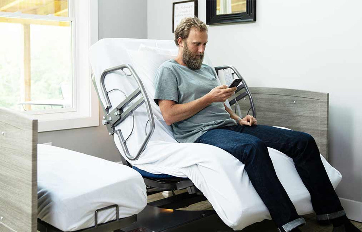 Hospital bed with raised head section and side rails, man sitting upright using a phone in a home bedroom setting