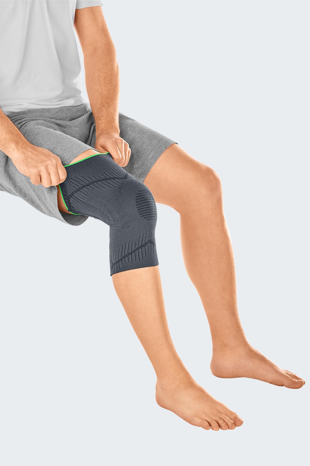 Knee support sleeve in gray knit fabric with green trim being put on by a seated man wearing gray shorts