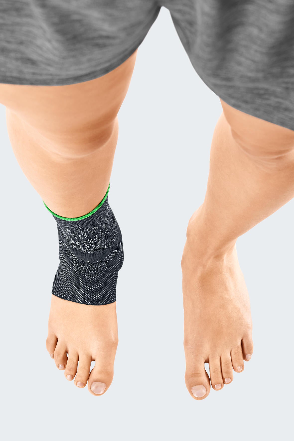 Ankle support sleeve in dark gray knit fabric with green trim band, worn on left ankle, open heel and toe design