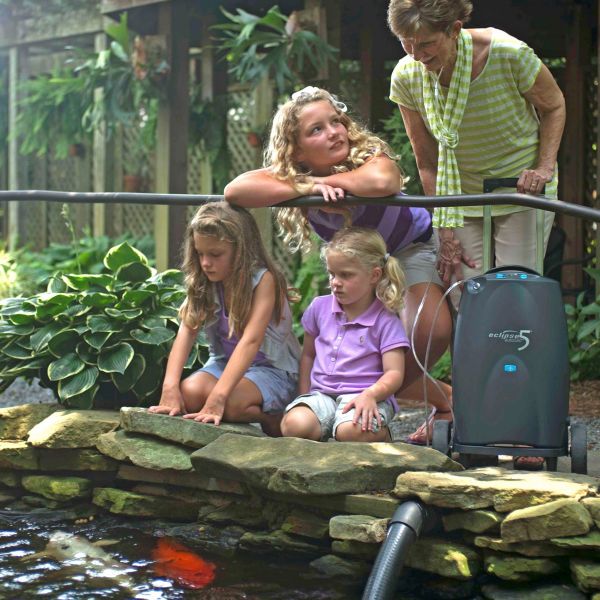 Portable oxygen concentrator on wheels beside a grandmother and three grandchildren watching koi fish at an outdoor pond