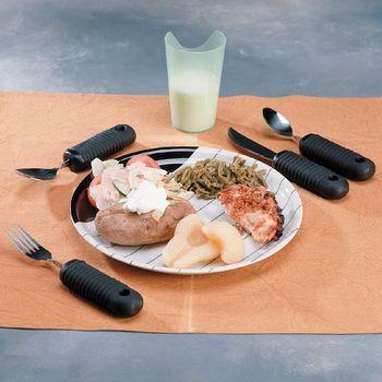 Weighted adaptive flatware set with black sure-grip handles arranged around a divided plate with food and glass of milk