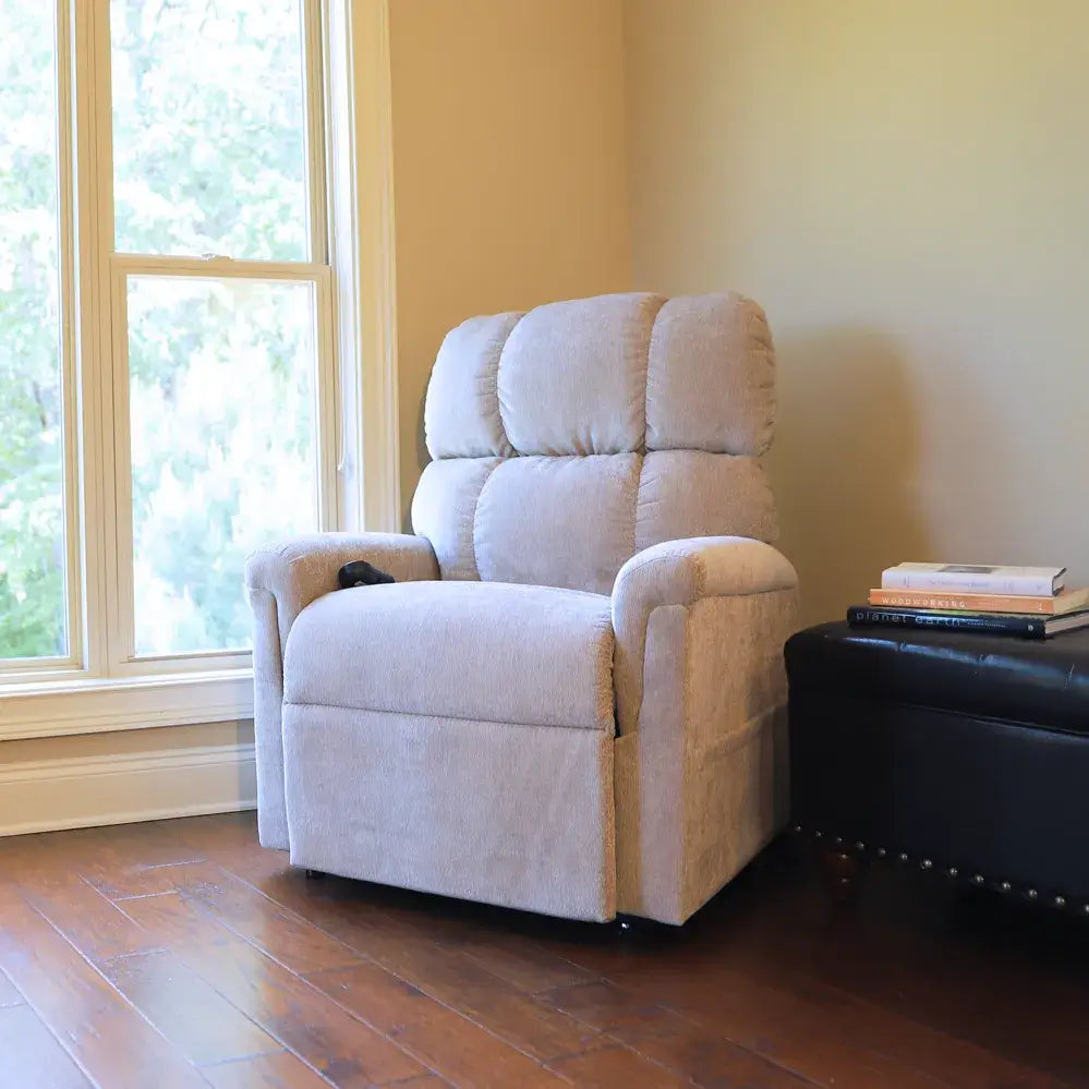 Power lift chair in light beige fabric with tufted high back, shown upright in a sunny home living room corner with hardwood