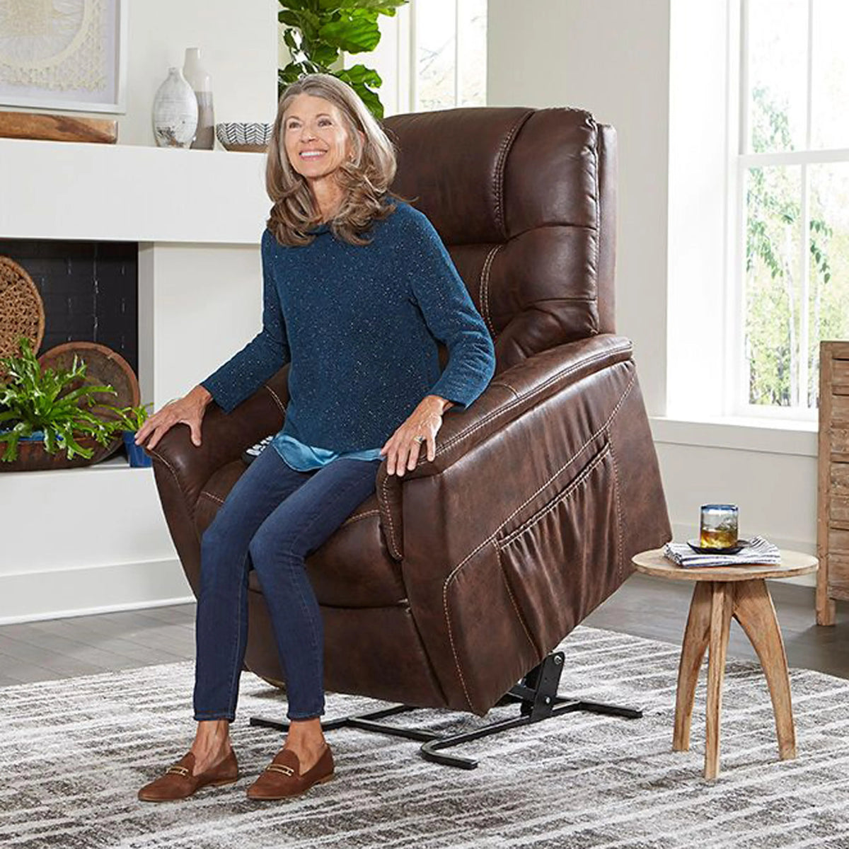 Power lift chair in brown faux leather shown in raised lift position with smiling older woman seated in living room setting