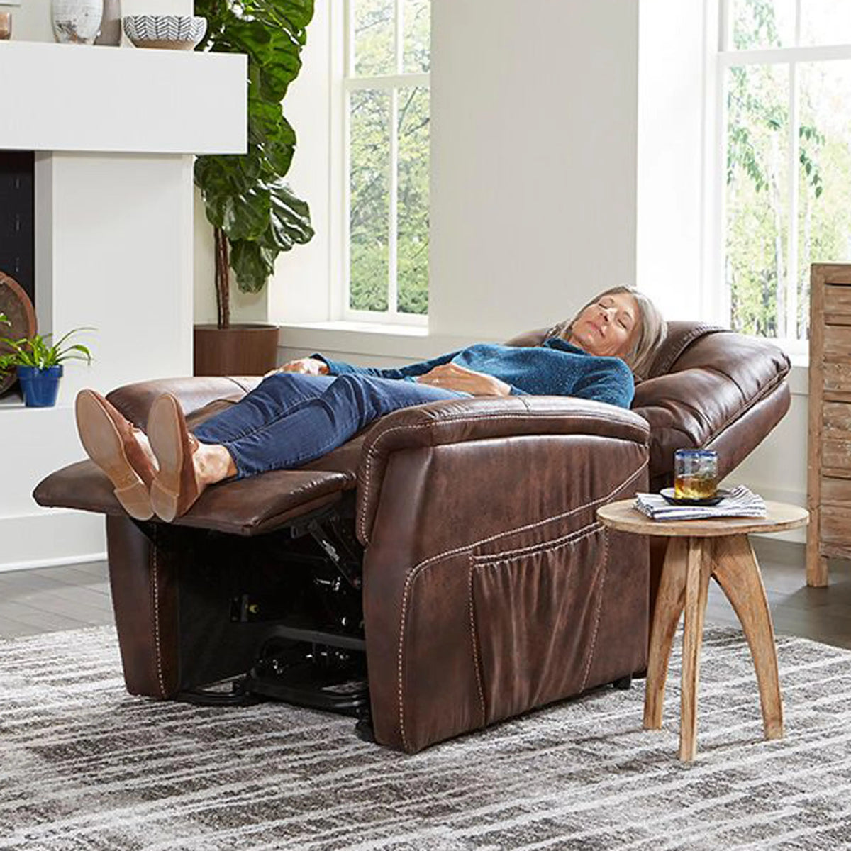 Power lift recliner chair in brown upholstery shown fully reclined with a woman resting in a living room setting