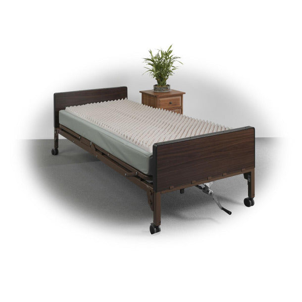 Convoluted foam pad on a hospital bed with dark wood side rails and casters, shown with a nightstand and plant nearby