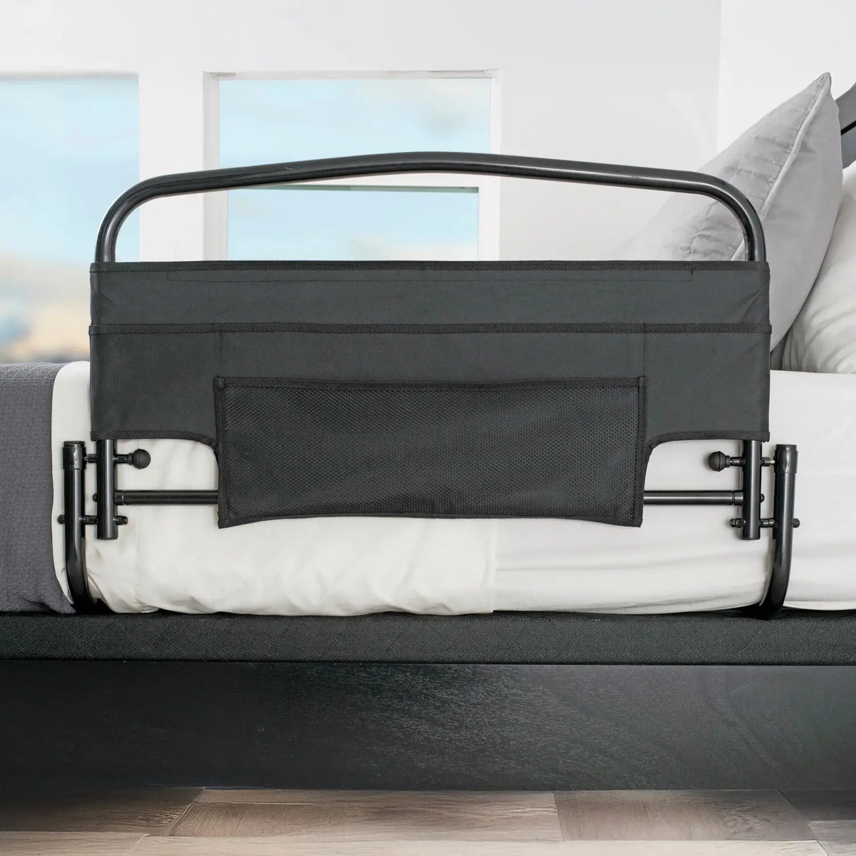 Bed rail with black padded fabric panel and mesh pocket, mounted on the side of a white mattress in a bedroom