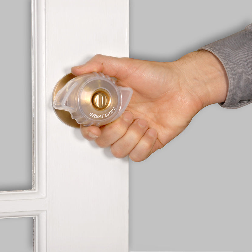 Door knob grip aid cover in clear plastic fitted over a brass knob, shown being turned by a hand on a white door