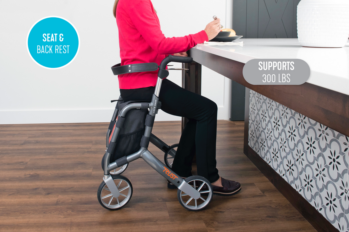 Rollator with seat and back rest supporting a seated woman at a kitchen counter, shown with large wheels and storage bag