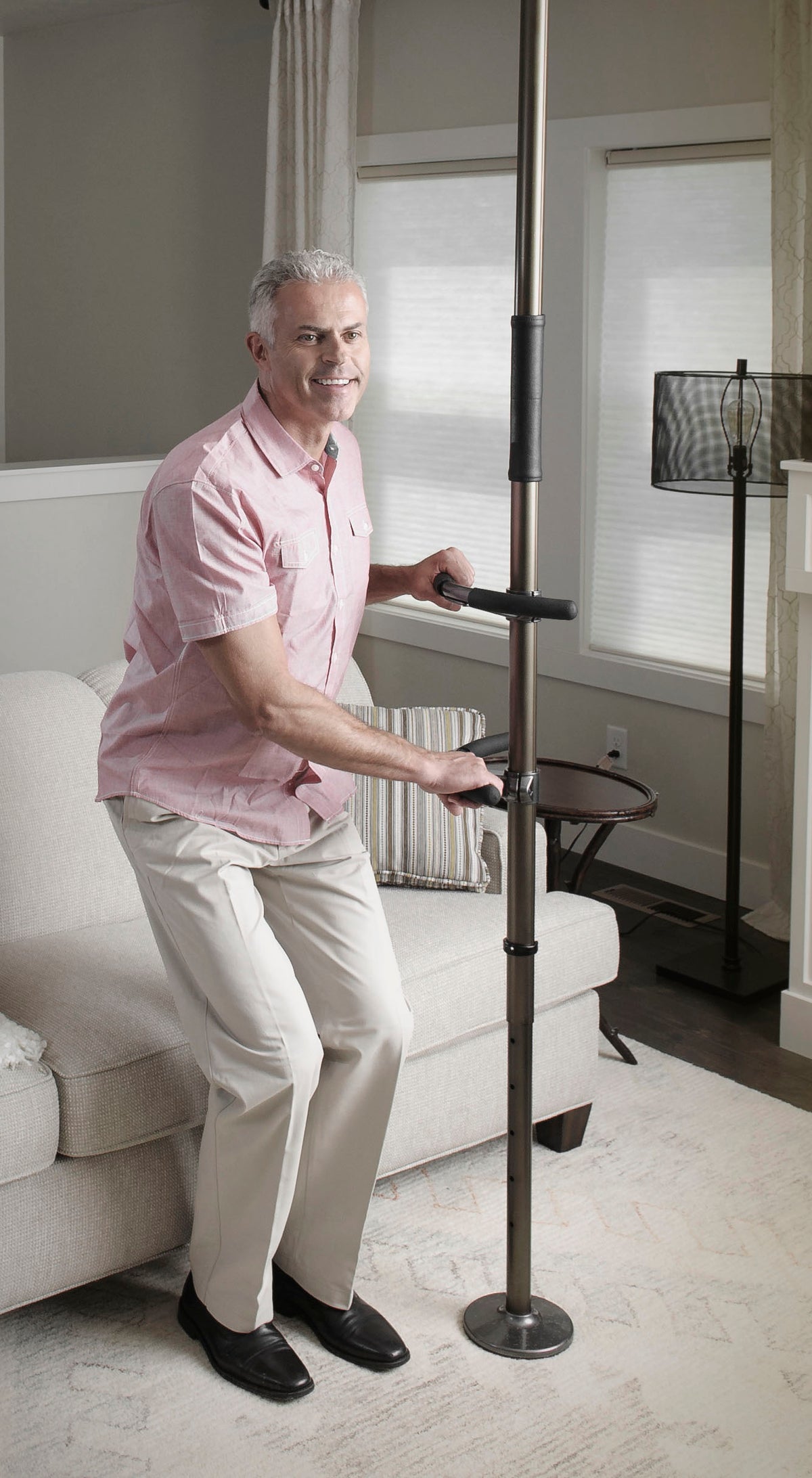 Floor-to-ceiling tension safety pole with two grab handles, used by a man rising from a living room sofa