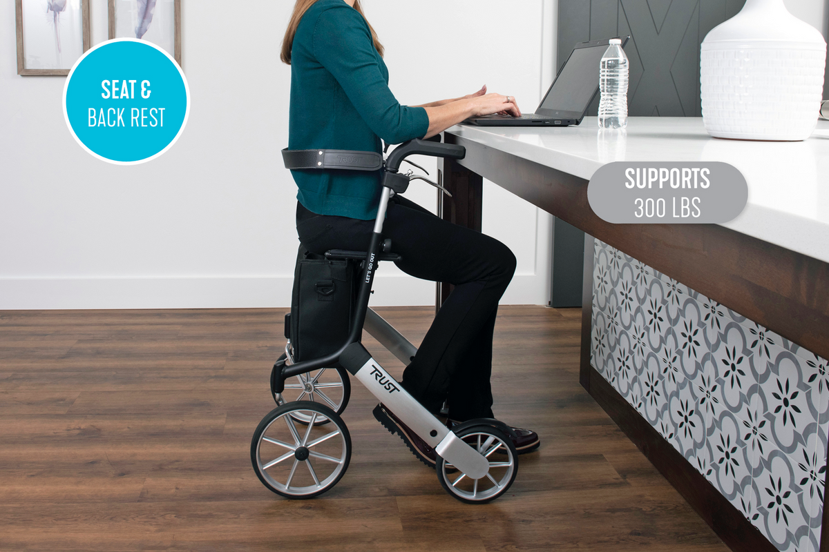 Rollator with seat and backrest used by a woman sitting while working on a laptop at a counter indoors