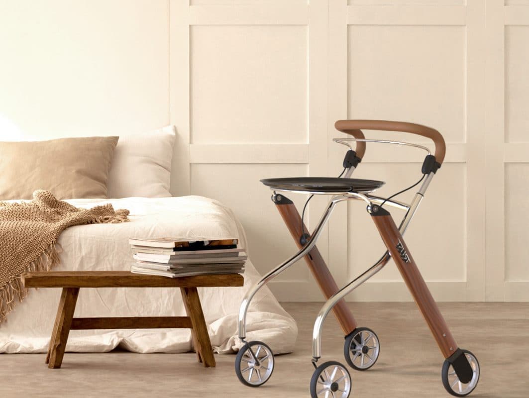 Indoor rollator with wood-tone frame, chrome accents, padded handles, and black tray, positioned beside a bed in a neutral