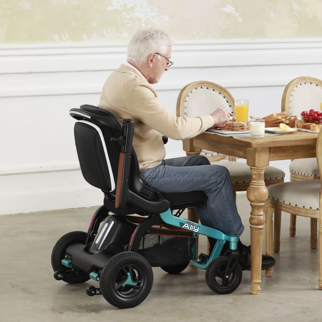 Power wheelchair in teal with elderly man seated at a dining table enjoying a meal indoors
