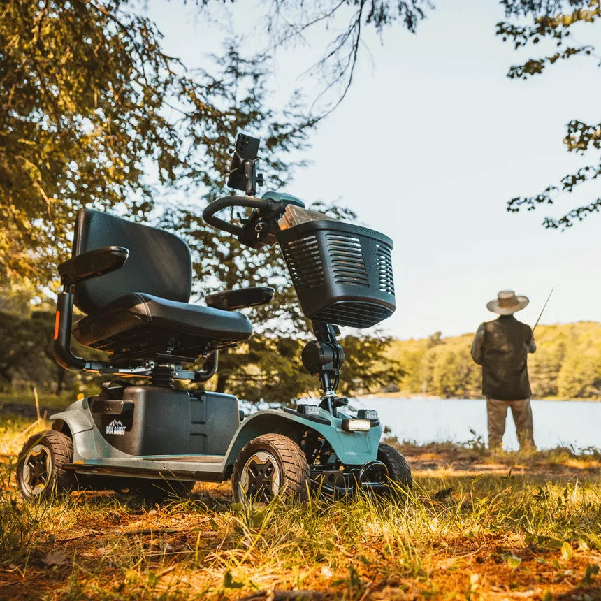 Power mobility scooter with front basket and rugged tires parked on grass near a lakeside, person fishing in background