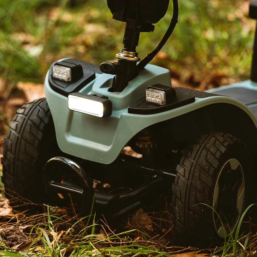 Power wheelchair front end with LED lights and rugged all-terrain tires, shown outdoors on grass and leaves