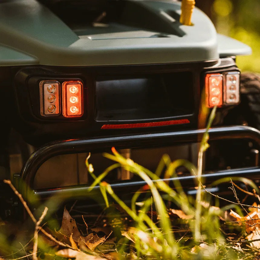 Power wheelchair rear panel with illuminated LED tail lights and black bumper guard, shown outdoors in grass and leaves