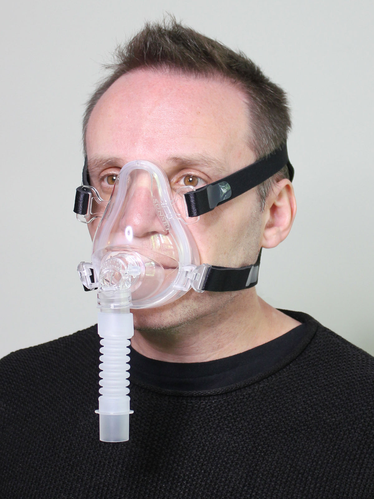 Full face CPAP mask with clear silicone cushion, black headgear straps, and corrugated elbow connector worn by a man