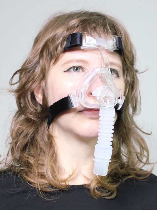 Nasal CPAP mask with clear silicone cushion, black headgear straps, and corrugated elbow connector worn by a woman