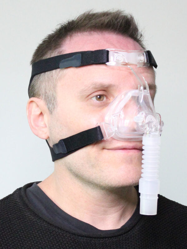 CPAP nasal mask with clear silicone cushion, black headgear straps, and corrugated elbow connector, worn by a man facing left