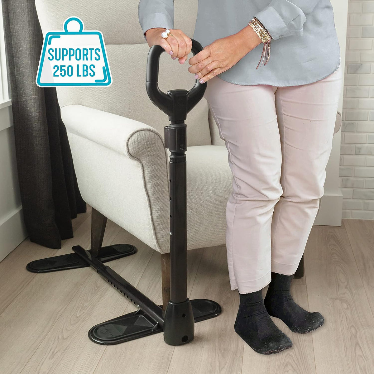 Couch cane with floor base and ergonomic handle being gripped by a woman standing beside a white armchair