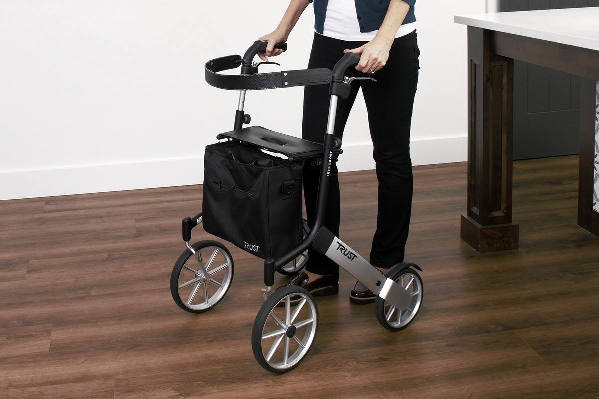 Rollator walker with four large wheels, fold-down seat, black storage bag, and tray, being used indoors by a person on