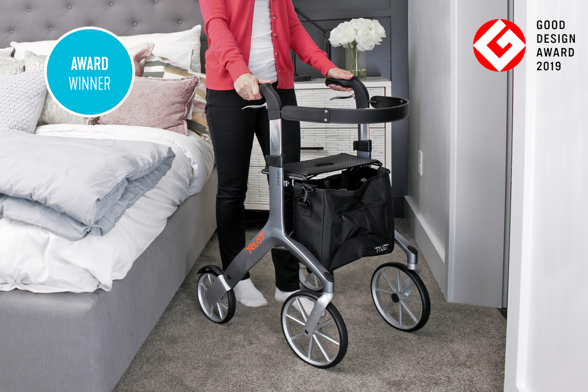 Lets Fly Rollator in silver with large wheels, seat, and black bag, used by a woman in a bedroom setting