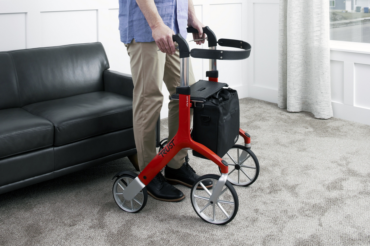Rollator walker in red with four large wheels, padded seat, hand brakes, and black bag, used indoors by a standing adult