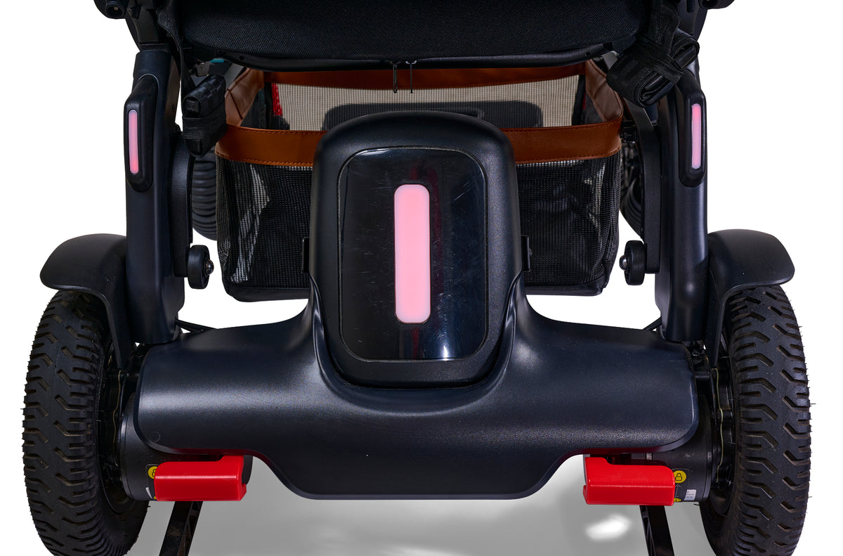 Power wheelchair rear view showing battery housing with pink LED indicator, rear wheels, side storage bags, and red safety