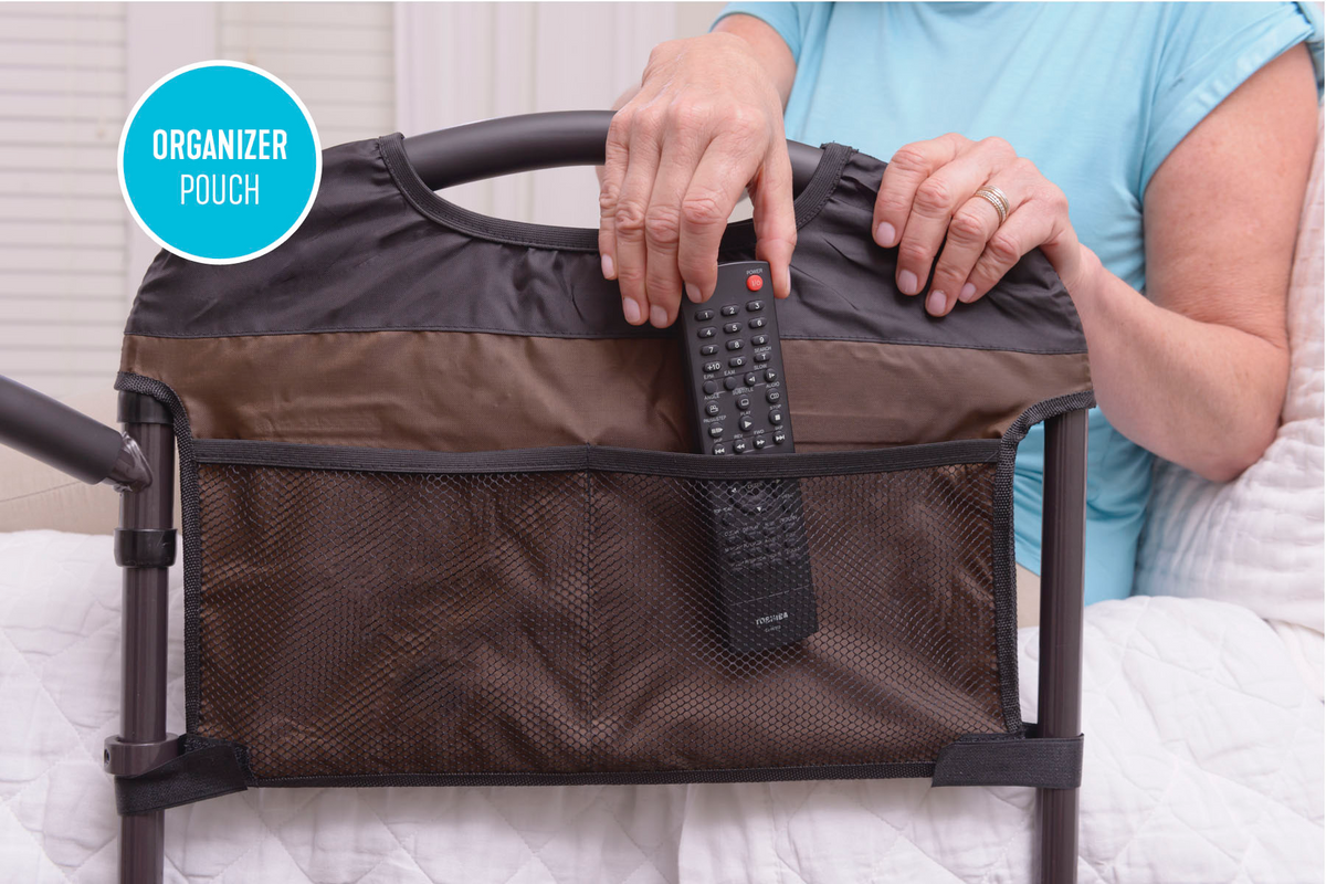 Bed rail organizer pouch with mesh pocket holding a TV remote, attached to a bed rail, shown in use by a seated adult