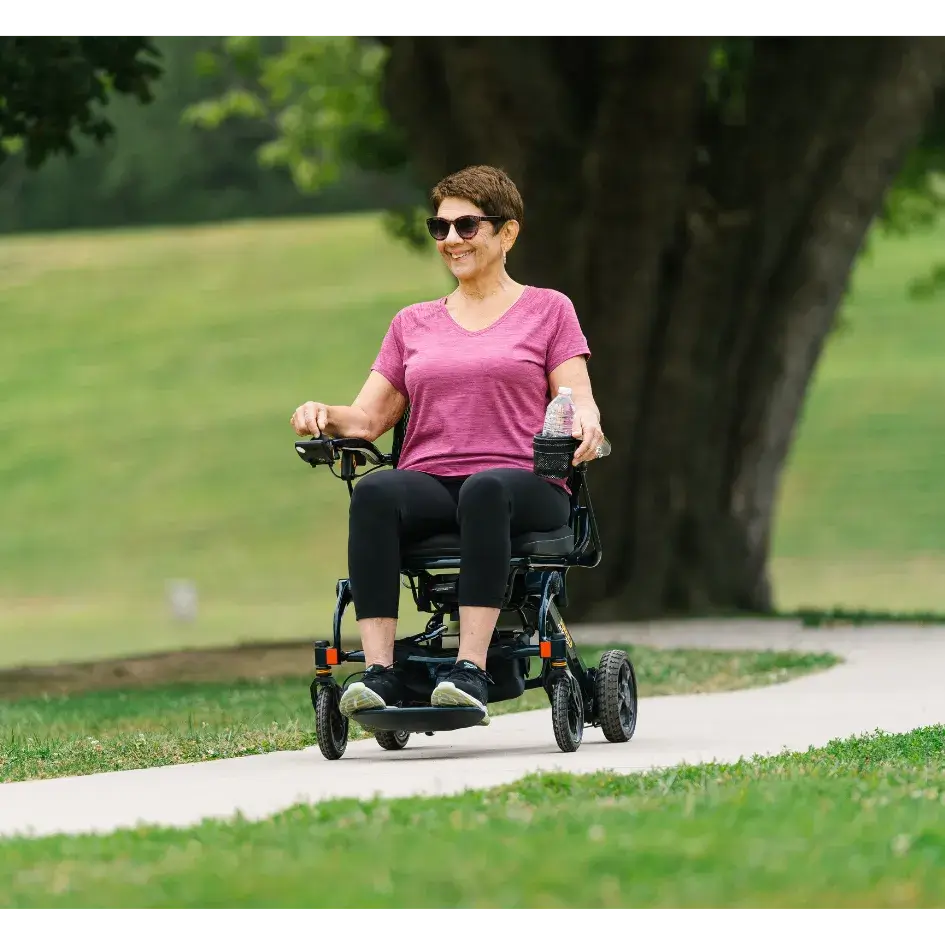 Power wheelchair carrying smiling woman along an outdoor park path, with cup holder and water bottle, green trees in