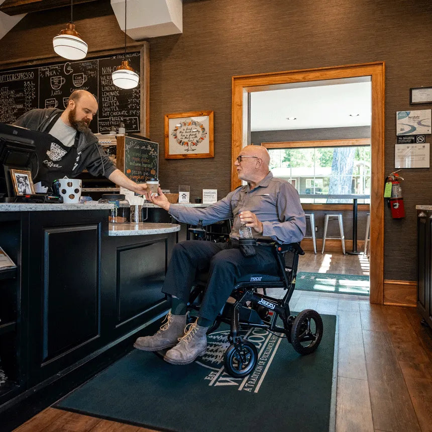 Power wheelchair user receiving a drink from a barista at a coffee shop counter while seated in a black Jazzy Ultra Light