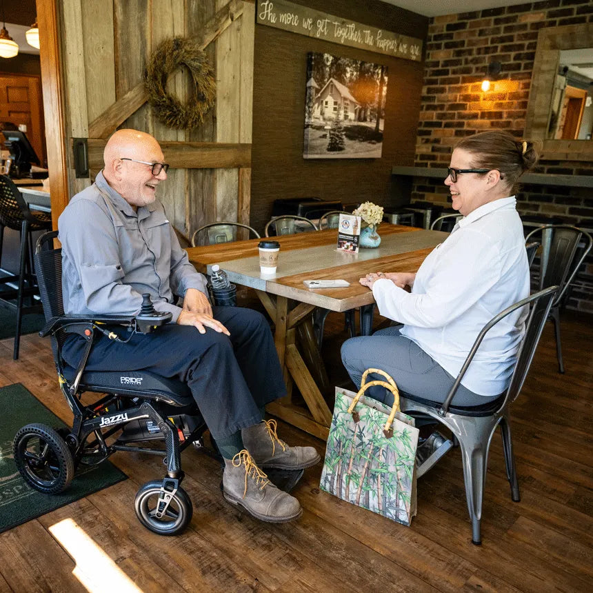Power wheelchair user and companion laughing together at a café table, with the Jazzy Ultra Light black folding power chair