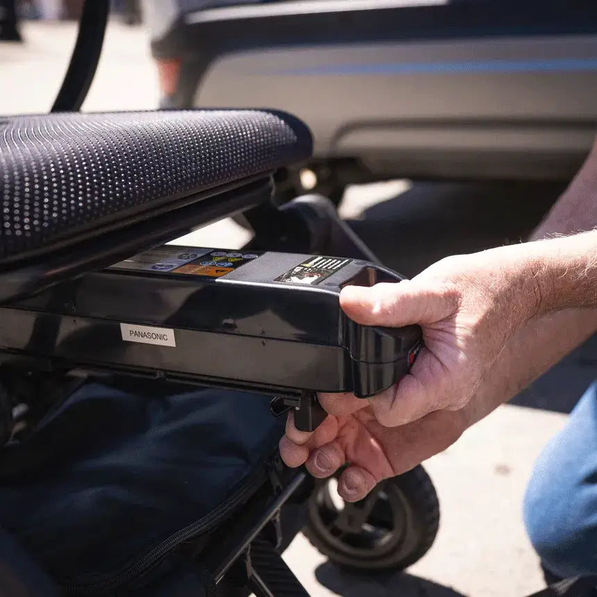 Power wheelchair battery pack being removed from beneath the seat of a Jazzy Rascal Carbon Cruiser by hand outdoors
