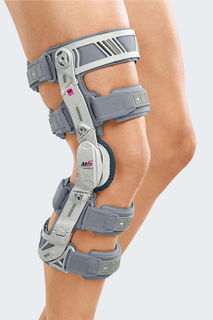 Knee brace with gray padded straps, hinged aluminum frame, and dial adjustment mechanism worn on a left leg, side view